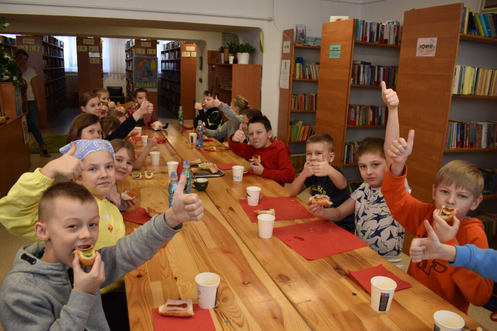 246 Dzieci siedzą przy stole i jedzą zrobione przez siebie kanapki. W tle regały z książkami