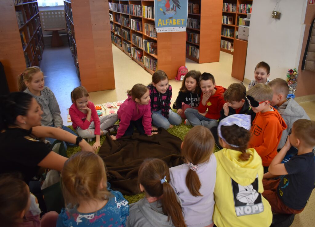 248 W Bibliotece na dywanie siedzą dzieci w kole i rozmawiają. W tle regały z książkami.