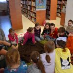 248 W Bibliotece na dywanie siedzą dzieci w kole i rozmawiają. W tle regały z książkami.