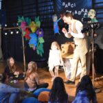 Na zdjęciu mężczyzna w garniturze przed dziećmi. Wygląda na to, że coś im przedstawia lub prezentuje.