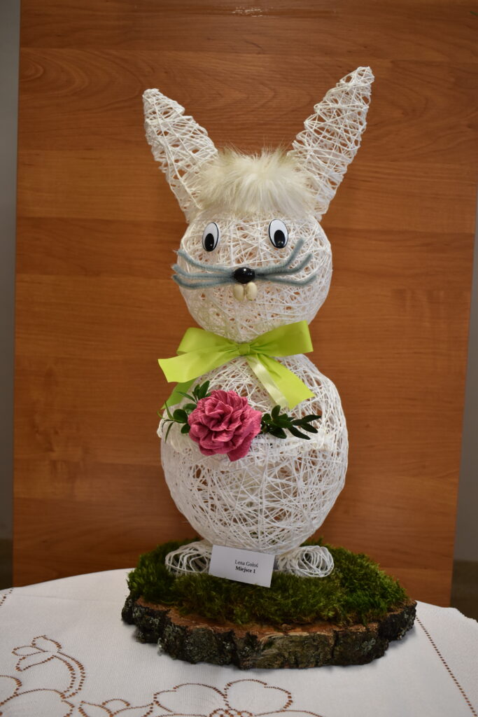 Na obrazku widnieje dekoracyjna rzeźba królika z muszką i okularami, która jest wystawiona na drewnianym pniu. Królik ma elegancki wygląd dzięki dodatkom, które ma na siebie.