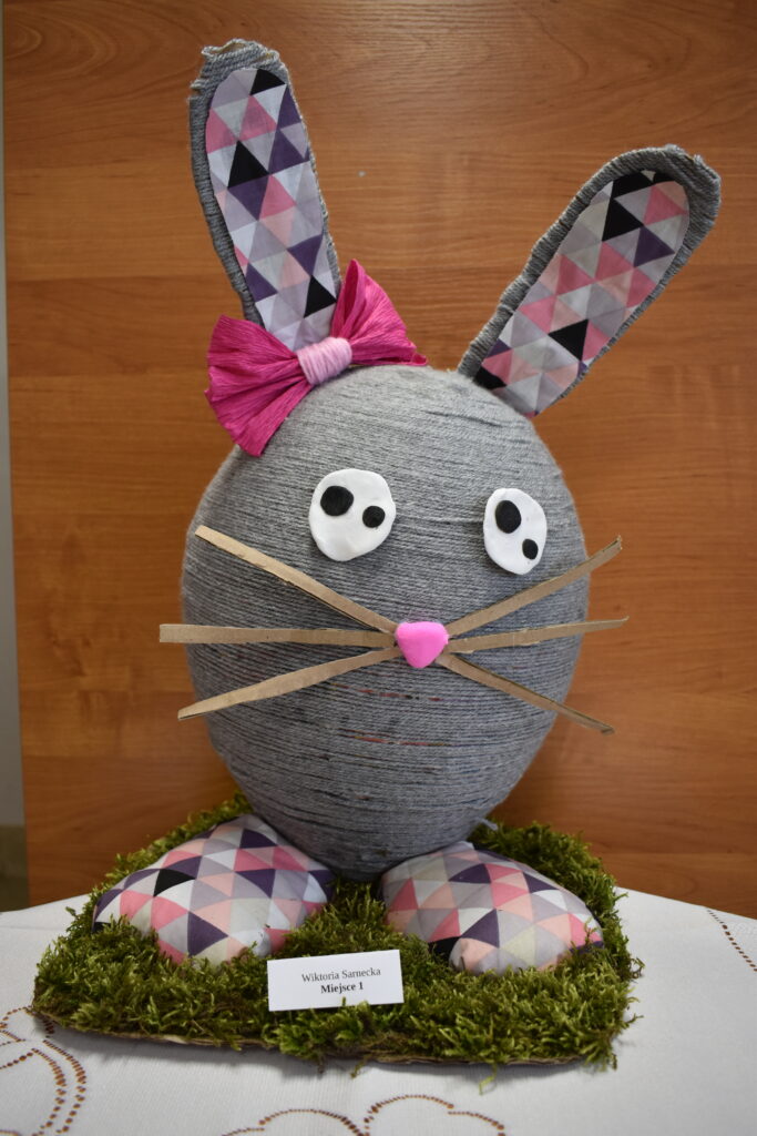 Na obrazku dekoracyjnym wielkiego królika, wykonanego z szarego i różowego materiału, który jest wystawiony na pościel z sztucznych trawy. Królik ma sympatyczną minę i brzmi na delikatnym w dotyku.