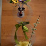 Obrazek przedstawia kreatywne, wiosenne kwiatowe z drucianą strukturą, którą znajdują się kwiatami, których centrum pojawia się fantazyjna twarz. Kompozycja uzupełniona jest gałązką utylizacyjną i prezentowaną na tle drewnianej powierzchni.