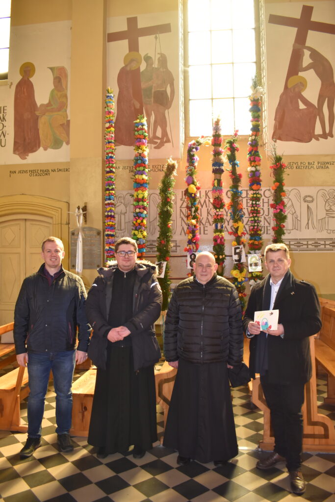 Na obrazku czterech mężczyzn stojących w kościele. W tle zaznaczamy dekoracje w postaci kolorowych wstążek.
