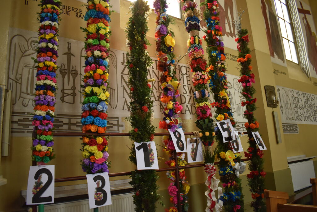 Na zdjęciu są kolorowe, ozdobnie przyozdobione palmy wielkanocne pionowo wystawione w kościele. Oznaczone z nich jest oznaczeniem, co wskazuje na udział w zawodach.