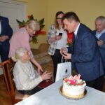 Na obrazku grupę osób świętujących urodziny starszej kobiety. Mężczyzna prezentuje tort siedzącej jubilatce.