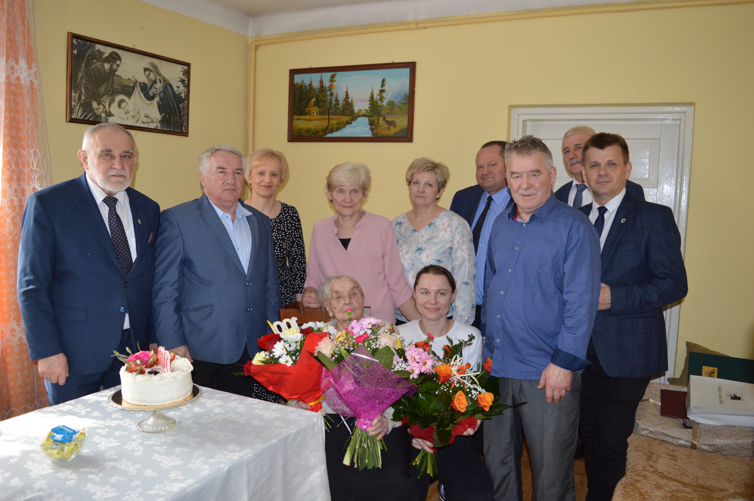 Na obrazku grupy dorosłych stojących razem w pomieszczeniu i uśmiechających się. Starsza osoba oraz młodszy dorosły dorosły przy stole, na którym znajduje się ciasto i kwiaty.