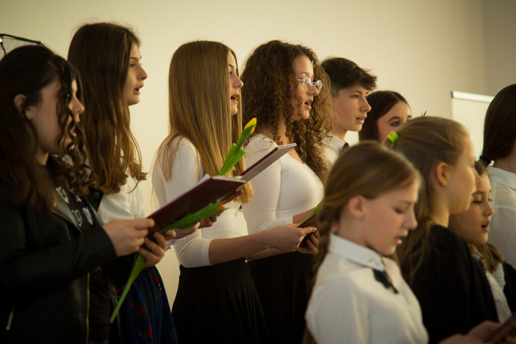 Na obrazku grupę dziewcząt, które śpiewają w chórze. Ubiorone są jednymi w jednolitych strojach, które są przeznaczone na dyrygencie, które prowadzą ich wykonanie.