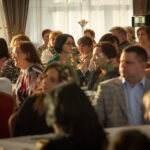 Na obrazku grupę osób siedzących przy stole, angażujących się w czasie. Wygląda na to, że można przystępować do posiłku, gdy na stole znajdują się różne naczynia i sztućce.