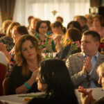 Na obrazku grupę ludzi siedzących wokół stołu, które entuzjastycznie oklaskują. Atmosfera jest radosna, a twarze uczestników wyrażają zadowolenie i aprobatę.