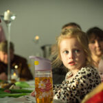 Na dane małej dziewczynki siedziba przy stole, na której znajdują się dane osobowe. Wokół niej ludzie, którzy uznają się za zaangażowani w jakieś zgromadzenie lub aktywność.