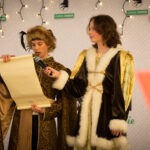 Na obrazku jest grupa ludzi, ubranych w stroje średniowieczne. Trzymają oni jakiś dokument lub kartkę papierową.