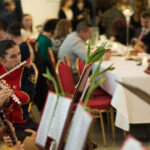 Na obrazku widać grupę ludzi, która siedzi przy stole. Wyglądają na to, że są zrelaksowani i prowadzą siebie nawzajem lub coś jedzą.