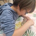 Na obrazku widzi młodą kobietę w okularach, która siedzi przy stole z tropikalnych liści, kształtuje glinę. Osoba ta jest skupiona na swojej pracy, a na niej rozmieszczone są różne narzędzia potrzebne do modelowania.