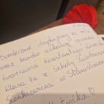 Na obrazku zbliżenie na notatkę na odręcznie po polsku na stronie zeszytu. Notatka udekorowano czerwonym płatkiem kwiatu.