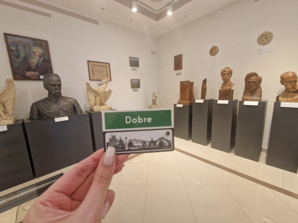 Na zdjęciu jest ręka trzymająca plakietkę z napisem „dobre” w muzeum popiersi i małe rzeźby przechowywane na postumentach. Kolekcja wydaje się być szczegółowa prezentowana, a osoba zawierająca informację o aplikacji jedno z usług.
