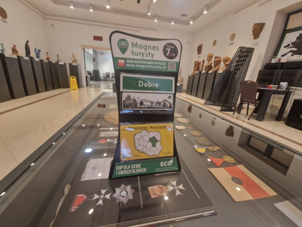 Na widocznym jest magnes turystyczny wystawiony na ladę w sklepie z pamiątkami w muzeum. W tle są różne broszury informacyjne i inne przedmioty.
