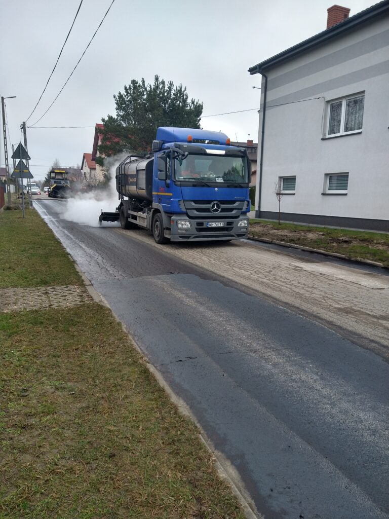 Na obrazku widocznym jest niebieska ciężarówka marki Mercedes, która spryskuje wodę w celu jej oczyszczenia. Na tle zdjęcia są inne ważne elementy konstrukcyjne oraz budynek, a wszystko to, co odbywa się podczas pochmurnego dnia.