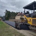 Na obrazku maszyny do układania nawierzchni marki Volvo, która wykłada świeży asfalt na drodze w rejonie mieszkalnym. W tle zauważamy domy oraz drzewa.