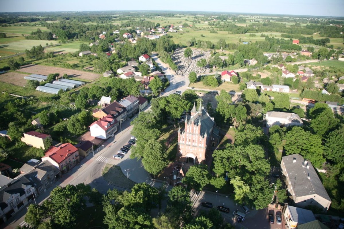 Na zdjęciu z lotem ptaka jest małe wiejskie miasteczko, w którym znajduje się obiekt, który jest symbolem innych budynków. Wokół miejscowości rozpościerają się zielone pola, podkreślając jej sielski charakter.