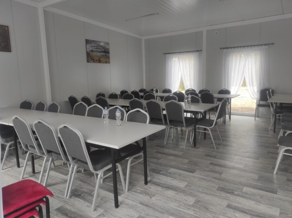 Obrazek przedstawia pustą salę konferencyjną z szarymi stołami i krzesłami na drewnianym wyposażeniu. Położenie jest jasne dzięki naturalnemu światłu wpadającemu przez zasłonięte okna.