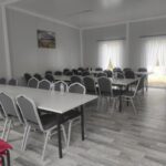 Obrazek przedstawia pustą salę konferencyjną z szarymi stołami i krzesłami na drewnianym wyposażeniu. Położenie jest jasne dzięki naturalnemu światłu wpadającemu przez zasłonięte okna.