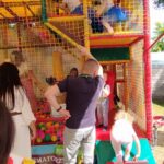 Dzieci bawią się na kolorowej konstrukcji placu zabaw z siatkami i miękkimi powierzchniami, gdy są dostępne w zasięgu sieci. Dziewczynka w niebieskiej sukience wspina się, a inne dziecko stoi na pochylni.