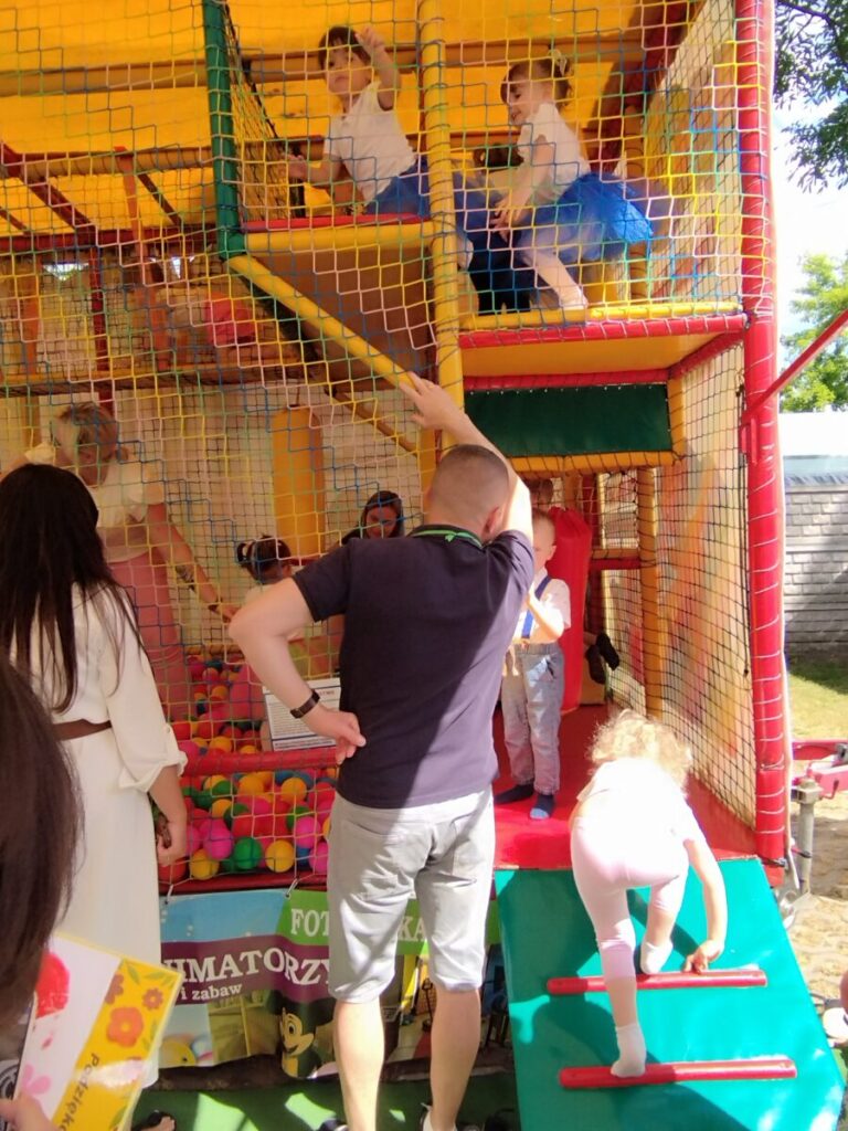Dzieci bawią się na kolorowej konstrukcji placu zabaw z siatkami i miękkimi powierzchniami, gdy są dostępne w zasięgu sieci. Dziewczynka w niebieskiej sukience wspina się, a inne dziecko stoi na pochylni.