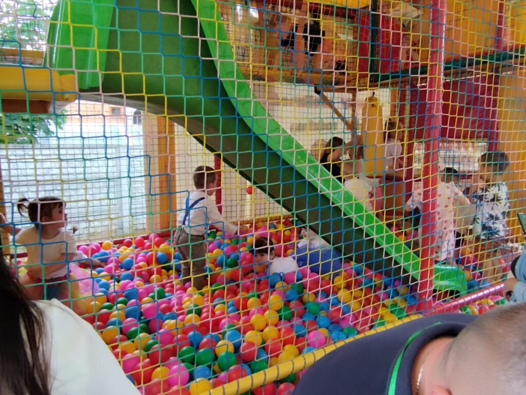 Na obrazku dzieci bawią się w gry z rzeczywistymi urządzeniami do gry w piłkę nożną, a nad nimi znajdują się zielona zjeżdżalnia. widoczna jest otoczona siatkowa konstrukcja wewnątrz sali zabaw.