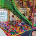 Na obrazku dzieci bawią się w gry z rzeczywistymi urządzeniami do gry w piłkę nożną, a nad nimi znajdują się zielona zjeżdżalnia. widoczna jest otoczona siatkowa konstrukcja wewnątrz sali zabaw.