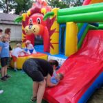 Na obrazku dzieci bawią się na dmuchanej zjeżdżalni ozdobionej motywami lwów, przy użyciu dorosłych. Kobieta pomaga jednemu z dzieci wspinać się na zjeżdżalnię, podczas gdy reszta czeka w pobliżu; całość odbywa się w plenerze.