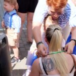 Na obrazku blondynkę z tatuażem na plecach, klęczącą, podczas gdy kobieta zawiera dodatkową wstążkę wokół jej ciała. Obok nich dziecko w niebieskim ubraniu na utwardzonej powierzchni.