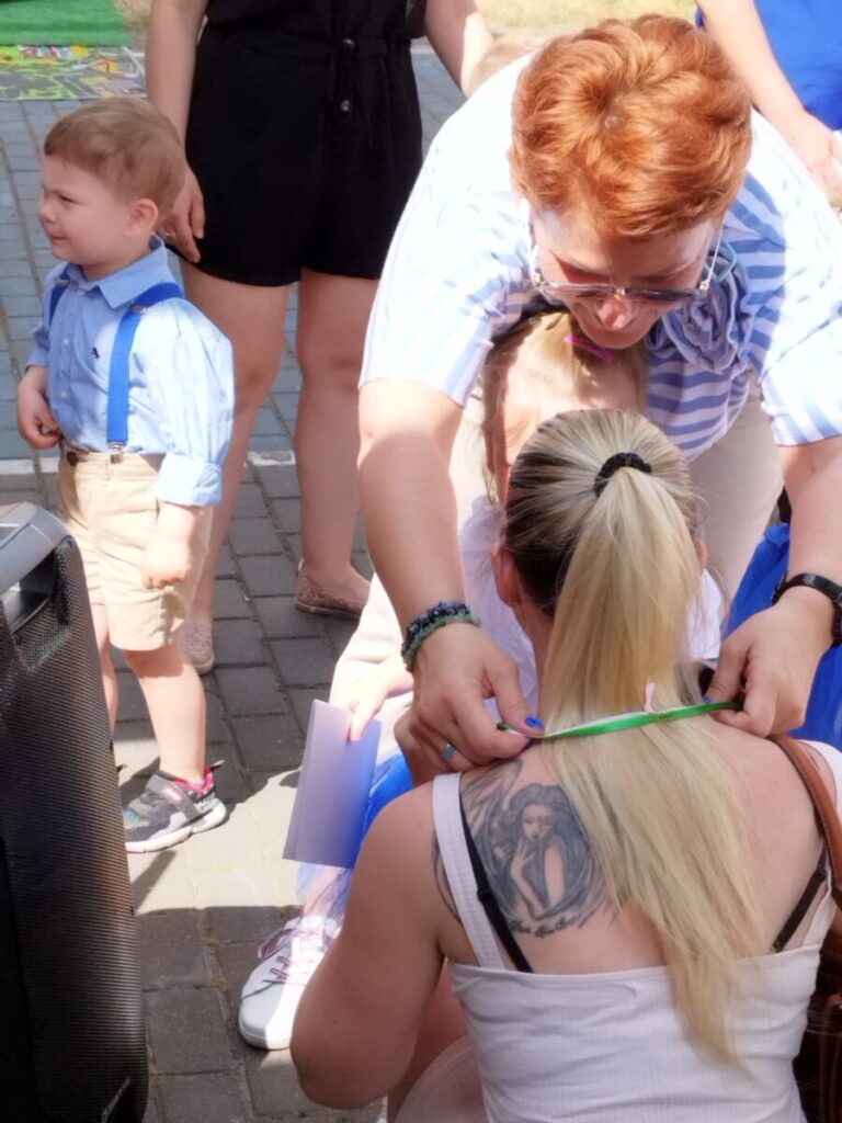 Na obrazku blondynkę z tatuażem na plecach, klęczącą, podczas gdy kobieta zawiera dodatkową wstążkę wokół jej ciała. Obok nich dziecko w niebieskim ubraniu na utwardzonej powierzchni.
