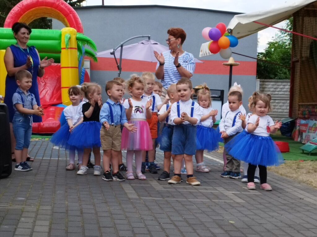 Na obrazku grupy małych dzieci, które jest dostępne na świeżym powietrzu przed dmuchanymi zamkami, otwierając coś przed nią. W tle dwie dorosłe osoby klaszczące, a wokół są balony i dekoracje.