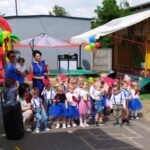 Na obrazku widać grupę małych dzieci przebranych w kostiumy, stojących na utwardzonym terenie przed dmuchaną zjeżdżalnią i kolorowymi namiotami, które są obsługiwane przez dorosłych. Wygląda na to, że dzieje się tam uroczyste spotkanie na świeżym powietrzu.