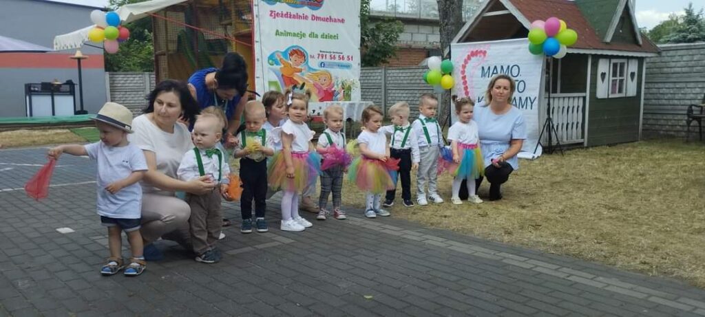 Na obrazku pojawia się grupa małych dzieci ubranych w kolorowe stroje, stojących i klęczących na otwartej, wybrukowanej przestrzeni. Towarzyszą im trzy osoby dorosłe, a w tle są baner i balony, co jest zawarte w nich lub w nich zawarte.
