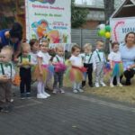 Na obrazku pojawia się grupa małych dzieci ubranych w kolorowe stroje, stojących i klęczących na otwartej, wybrukowanej przestrzeni. Towarzyszą im trzy osoby dorosłe, a w tle są baner i balony, co jest zawarte w nich lub w nich zawarte.