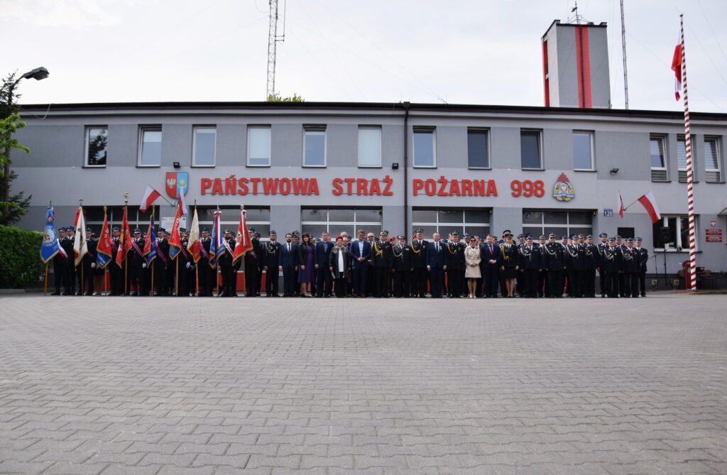 Na obrazku występuje duża grupa ludzi w mundurach i formalnych strojach, stojących przed remizą strażacką, obok której powiewają różne flagi. Budynek ma znak wskazujący na Państwową Straż Pożarną i numer alarmowy 998.
