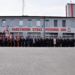 Na obrazku występuje duża grupa ludzi w mundurach i formalnych strojach, stojących przed remizą strażacką, obok której powiewają różne flagi. Budynek ma znak wskazujący na Państwową Straż Pożarną i numer alarmowy 998.