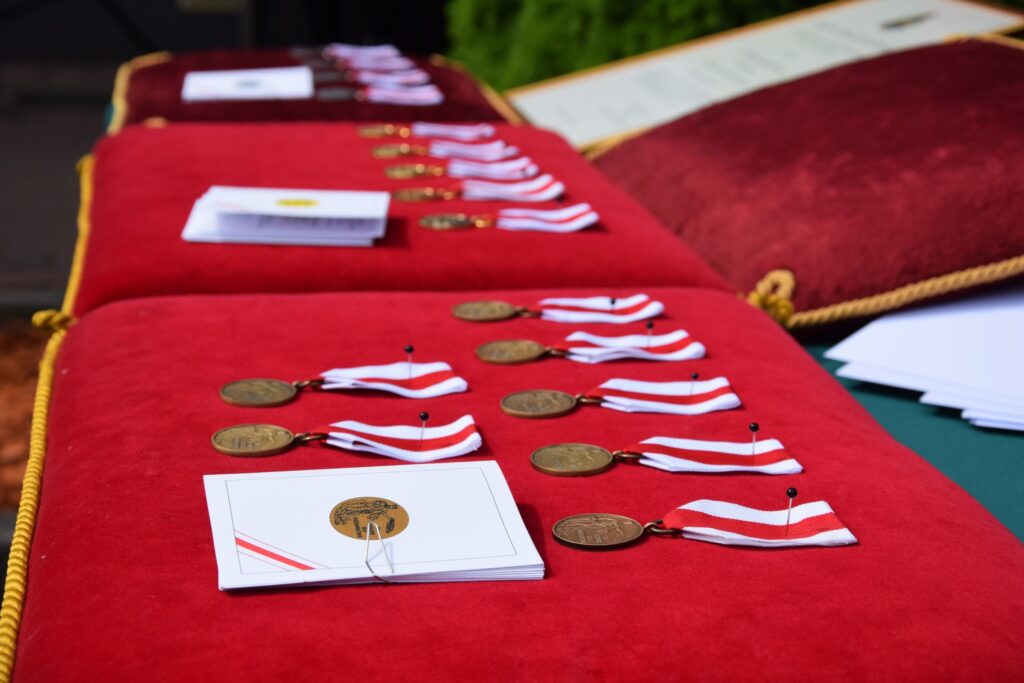 Na widocznym tle jest zestaw medali z czerwono-białymi wstążkami oraz certyfikaty, elegancko ułożony na czerwonym aksamitem. Wygląda na przygotowanie do ceremonii rozdania nagród lub wyróżnień.