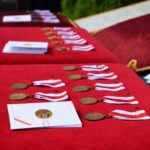 Na widocznym tle jest zestaw medali z czerwono-białymi wstążkami oraz certyfikaty, elegancko ułożony na czerwonym aksamitem. Wygląda na przygotowanie do ceremonii rozdania nagród lub wyróżnień.