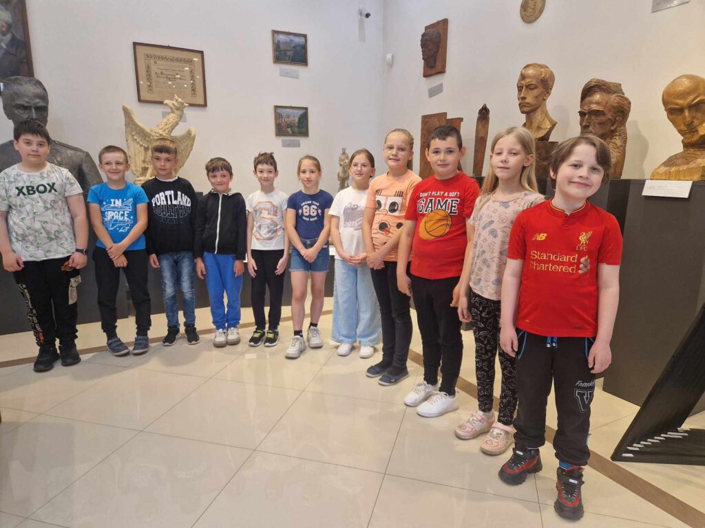 Na obrazku grupa jedenastu dzieci stoi w galerii muzealnej, równo ustawionej obok siebie. W tle widoczne są popiersia oraz obrazy w ramach, które zdobią ścianę sali.