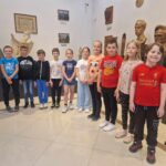 Na obrazku grupa jedenastu dzieci stoi w galerii muzealnej, równo ustawionej obok siebie. W tle widoczne są popiersia oraz obrazy w ramach, które zdobią ścianę sali.