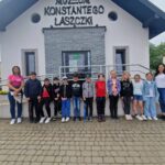 Na obrazku widać grupę dzieci i dwóch dorosłych stojących przed widocznym oznaczonym napisem „Muzeum Konstantego Laszczki”. Tło budynku fizycznego, że jest miejscem kultury, może oferować wycieczki edukacyjne lub wystawy związane z życiem i twórczością artysty.