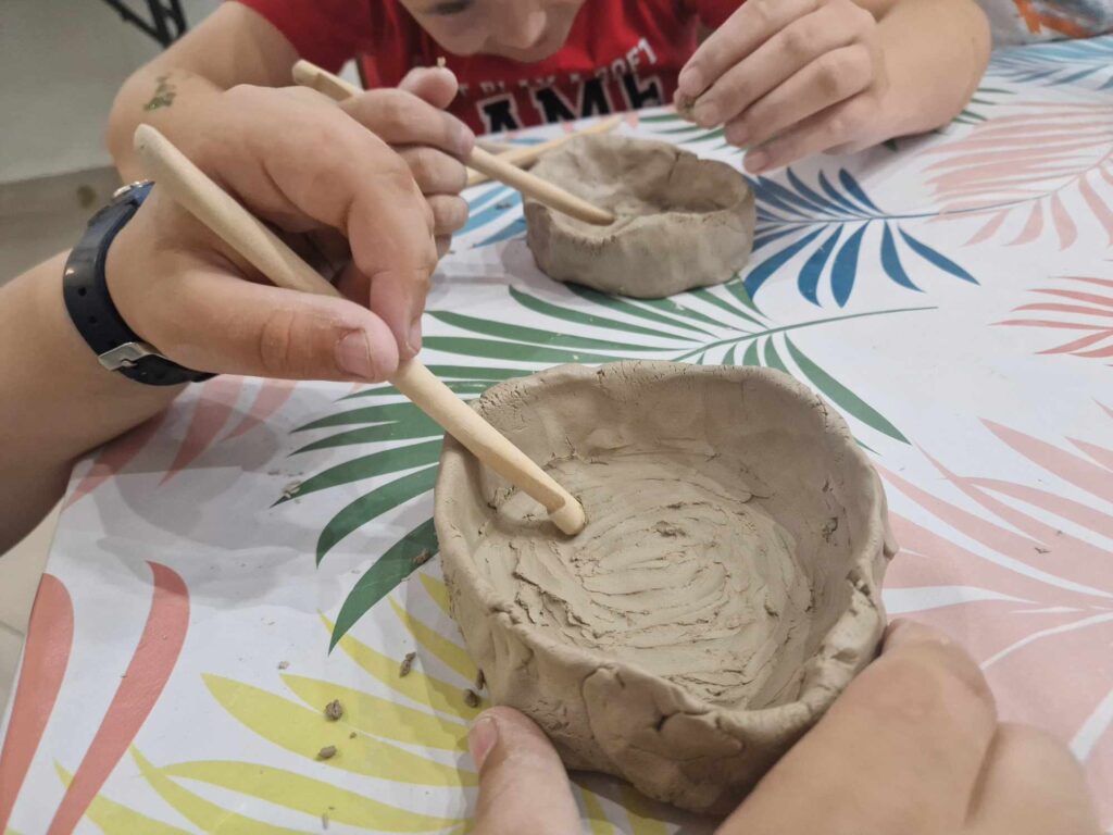 Na obrazku dwóch dzieci modeluje się gliniane miski przy użyciu narzędzia drewnianego, pochylając się nad kolorowym obrusem ozdobionym wzorami liści. występuje ich skupienie i precyzję, co charakterystyczna kreatywna i angażująca zabawę.
