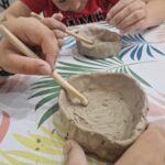 Na obrazku dwóch dzieci modeluje się gliniane miski przy użyciu narzędzia drewnianego, pochylając się nad kolorowym obrusem ozdobionym wzorami liści. występuje ich skupienie i precyzję, co charakterystyczna kreatywna i angażująca zabawę.