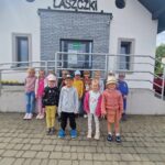 Grupa dzieci stoi przed wejściem do Muzeum Konstantego Laszczki, pozując do zdjęć. Są ubrani w codzienne stroje i czapki, a nad widocznym jest znak muzeum.