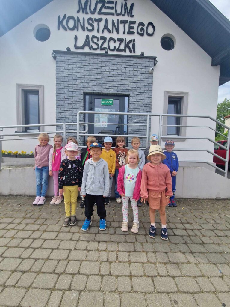 Grupa dzieci stoi przed wejściem do Muzeum Konstantego Laszczki, pozując do zdjęć. Są ubrani w codzienne stroje i czapki, a nad widocznym jest znak muzeum.