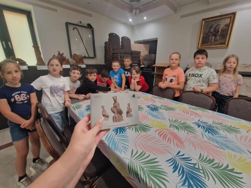 Na obrazku widocznym grupy dzieci stojących wokół stołu z kolorowym obrusem. Na pierwszym planie trzyma broszurę, na której znajdują się zdjęcia.