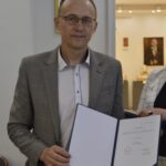 Na obrazku mężczyzn w okularach i w kratkowanym marynarce, który trzyma otwarty segregator. Scena rozgrywająca się w pomieszczeniu.